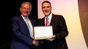 Two men posing together for a photo, shaking hands and holding a BiB award certificate