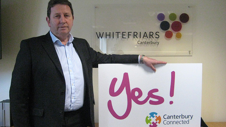 A man outside Whitefriars Canterbury holding a sign that says Yes! Canterbury Connected BID