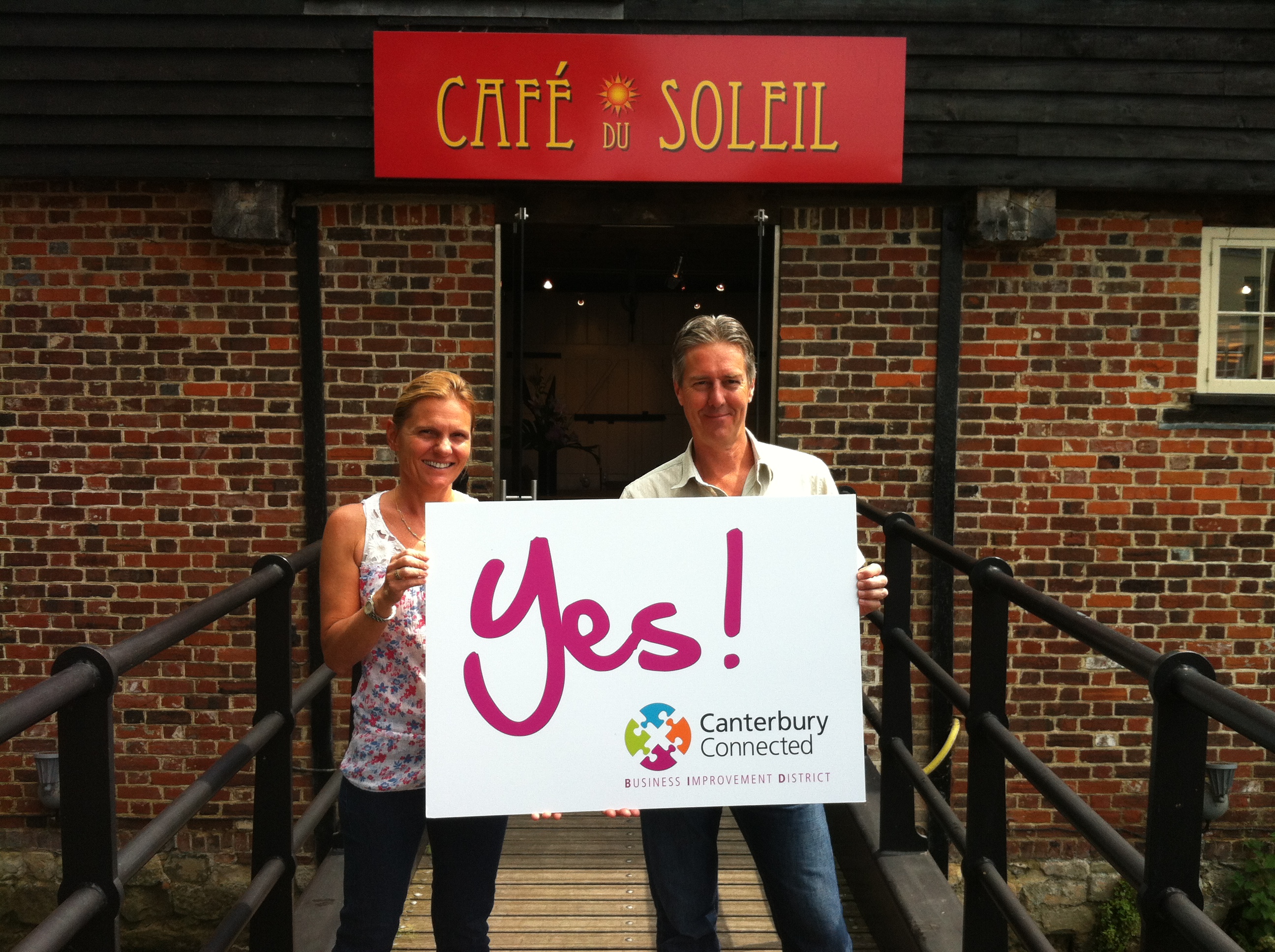 A man and a woman outside Cafe Du Soleil holding a sign that says Yes! Canterbury Connected BID
