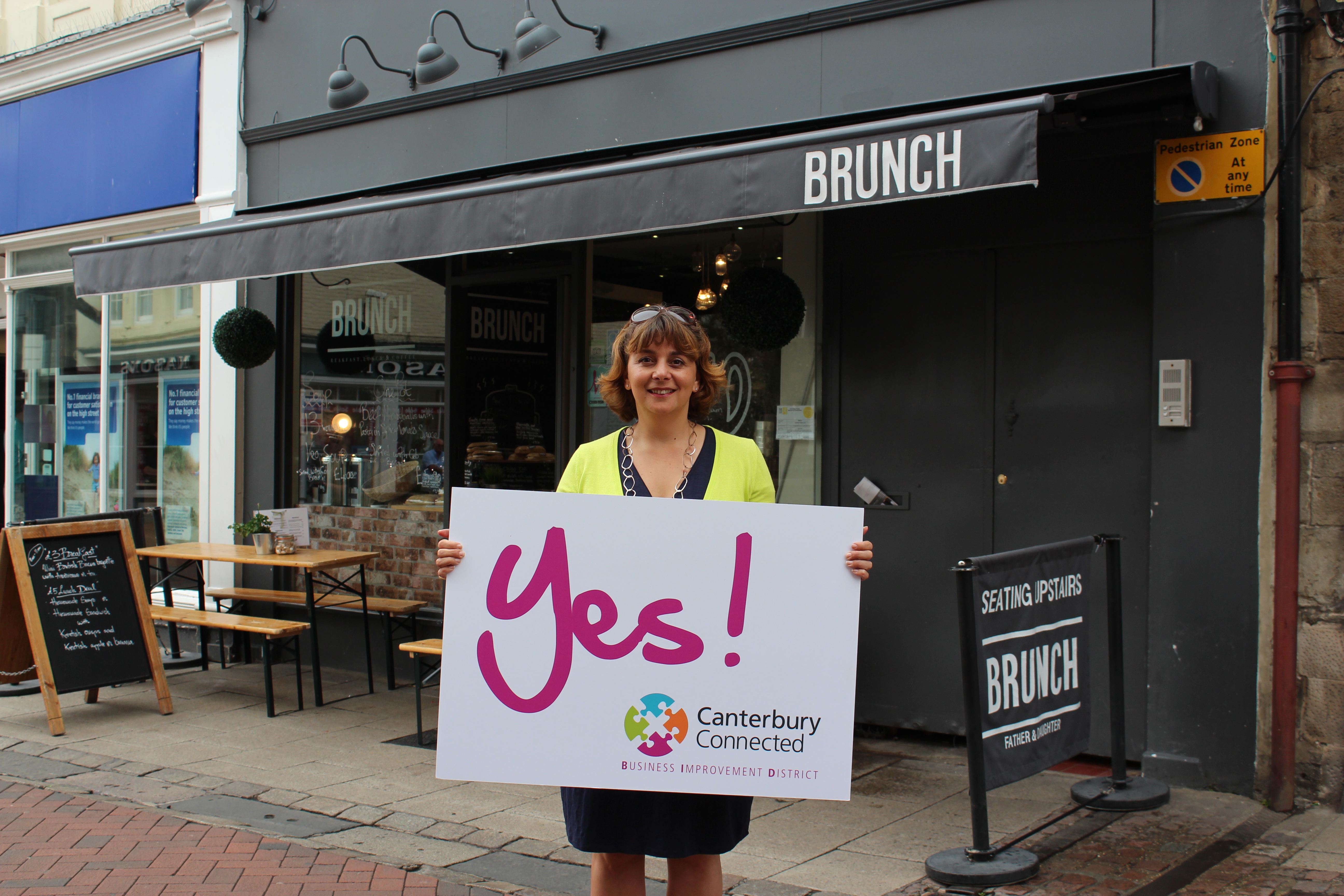 A woman outside Brunch holding a sign that says Yes! Canterbury Connected BID