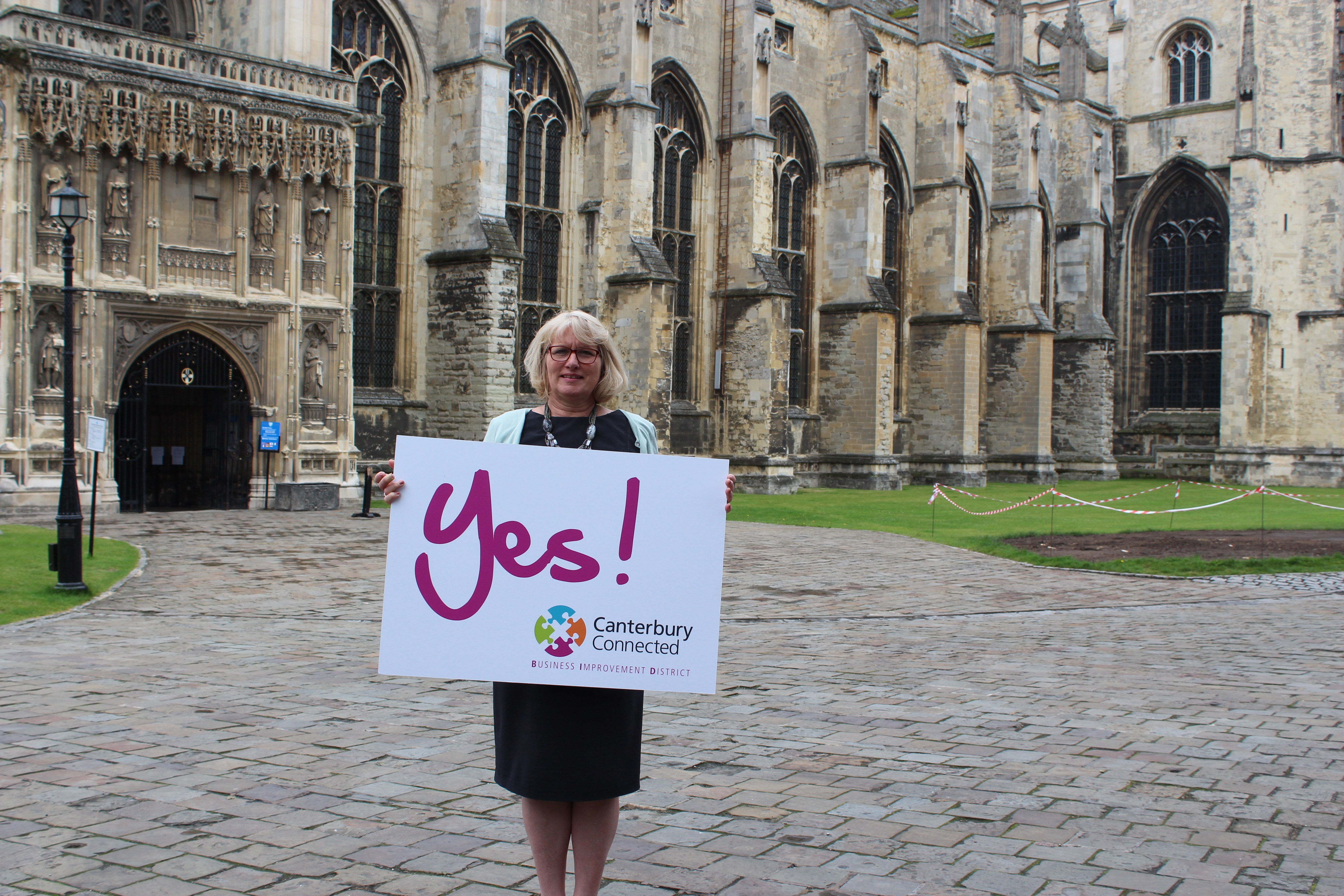 A woman outside Canterbury Cathedral holding a sign that says Yes! Canterbury Connected BID