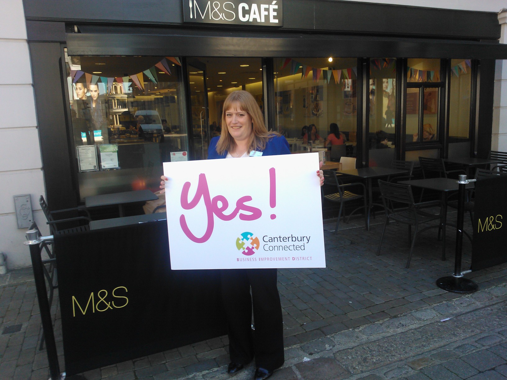 A woman outside M&S Cafe holding a sign that says Yes! Canterbury Connected BID