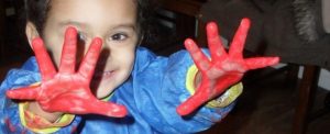 A small child holding out her hands in glee, covered in red paint
