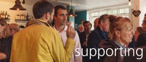 A photo of a room of people socialising with the word supporting over it in white text