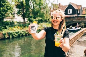 A photo of a woman by a riverside smiling and holding up two City Feast drinks