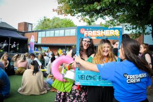 A photo of two female students at a UCA freshers event, smiling and standing in a cut out photo frame