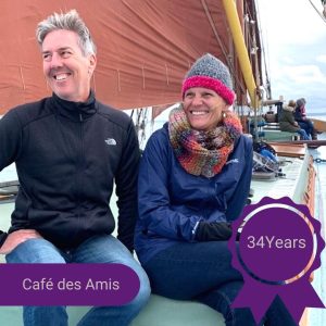 A photo of a man and a woman sitting on a boat and smiling, with text over it that reads 'Café des Amis - 34 years'